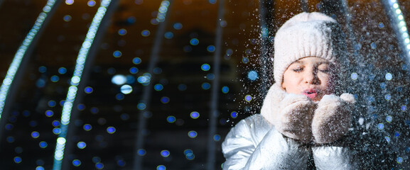 Girl and snow. Girl blowing on palms with snow at night with blue backlight and blurry lights. Banner