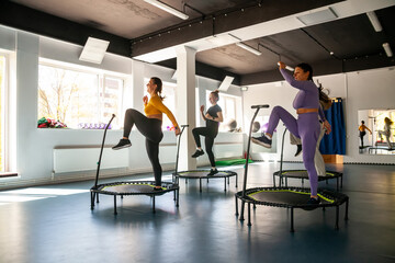 Group of happy women warming up before training in   gym jumping on   trampoline - obrazy, fototapety, plakaty