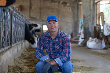 A contented man sits next to cows on a farm.