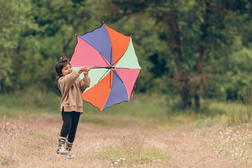 A little preschooler girl in a beige sweatshirt raises a multi-colored umbrella up to the sky in nature, she smiles and is happy, free