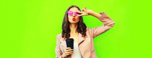 Obraz na płótnie Canvas Portrait of beautiful young woman blowing her lips sending air kiss posing wearing pink sunglasses, leather jacket on green background