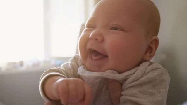 Cute newborn girl with plump cheeks smiles widely feeling happily at home. Mother holds baby with hands against bright window in bedroom close view