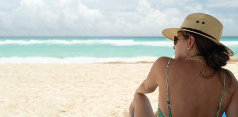 young hispanic woman with sunglasses and hat sitting looking at the sea