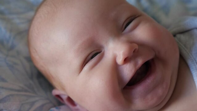 Adorable little girl with plump cheeks smiles wide lying on soft bed at home. Portrait of sweet newborn baby in bodysuit feeling happily closeup