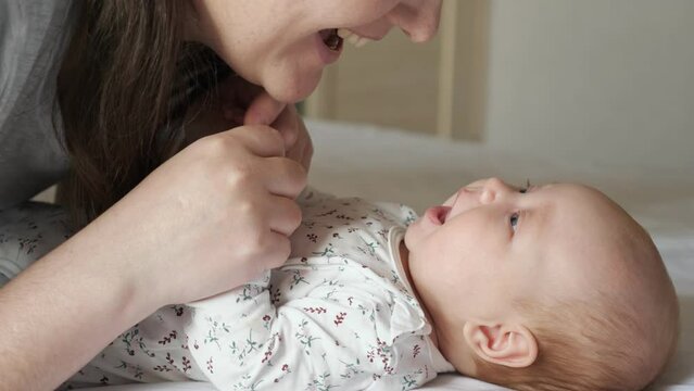 Happy mother kisses plump cheeks of newborn daughter lying on soft bed at home. Brunette woman spends time with baby girl in bedroom, close up