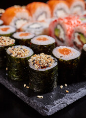 Close up of rolls with sauce and sesame seeds on a black flat plate.
