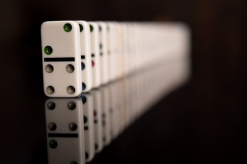 White domino bones stand on a black mirror surface. Domino effect. Domino effect and chain reaction concept. The background is blurry.