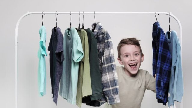 The little boy appears from behind the clothes rack. The boy looks through his shirts and T-shirts hanging on hangers.