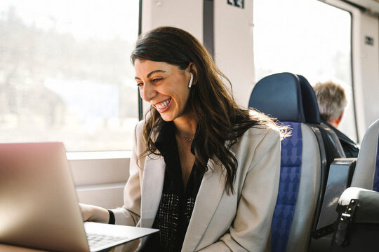 Happy businesswoman with in-ear headphones doing video call through laptop in train