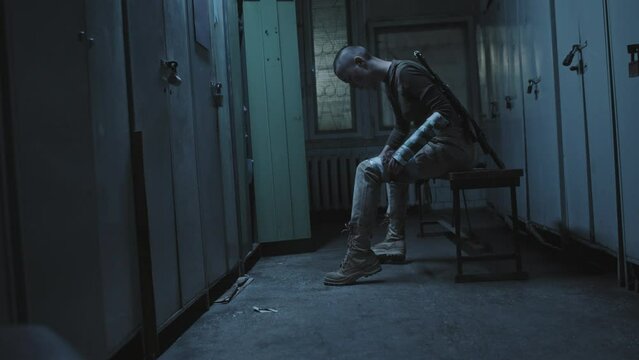 Wide side view of injured military woman with shaved head sitting in abandoned dark locker room, patching up wounds on leg with duct tape