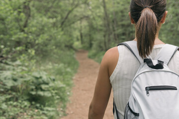 Woman walking along the path near the river. Young girl with backpack and white t-shirt wandering through the middle of the leafy path of the forest.