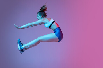 Moments. Young girl, female athlete training long jump technique isolated on pink studio background with blue neon filter, light. Concept of action, motion, speed