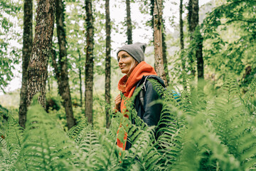 Young active woman hiker in a jacket and a hat with a backpack in a thicket of ferns in the forest