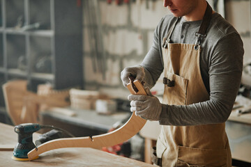 Moody closeup of young carpenter sanding wood while building handcrafted furniture in workshop,...