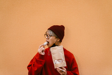 Asian boy wearing eyeglasses eating cookie while standing by wall