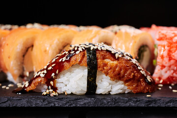 One piece of Nigiri sushi with smoked salmon, soy sauce and sesame seeds on a slate of black stone.