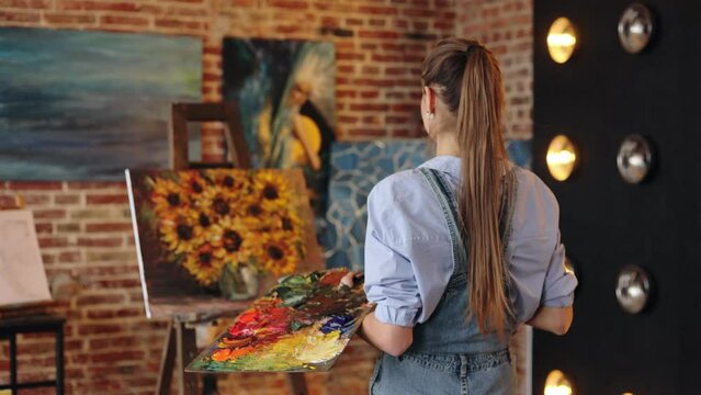 Young beautiful female artist painting still life with sunflower on canvas using oil paintings and palette knife. Painter creating artwork in art studio. Relaxation, leisure, hobby, stress management.