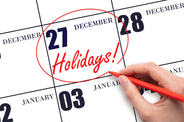 Hand drawing a red circle and writing the text Holidays on the calendar date 27 December. Important date.