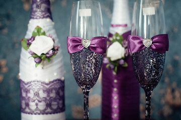 A pair of decorative purple champagne bottles and glasses for the bride and groom stand in a room, against a background of blue wallpaper.