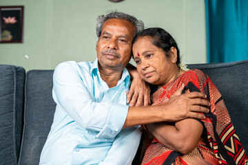 Senior man consoles by embracing his thouhgtful wife at home while on sitting sofa - concept of family, emotional love and support