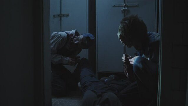 Low angle of two creepy zombies feeding on dead body on floor in dark abandoned building, then spotting and following invisible person who holding flashlight