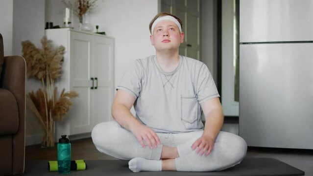 The guy stretches his neck while sitting on the floor in the lotus position at home in the living room in sportswear. Sport