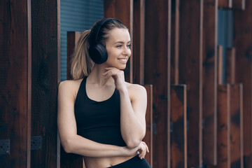 Blonde girl leaning on woden wall, listening music in earphones.
