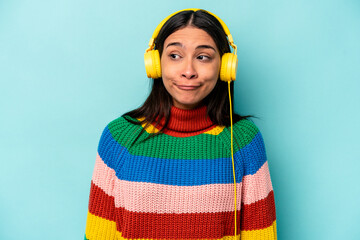 Young hispanic woman listening to music isolated on blue background shrugs shoulders and open eyes...