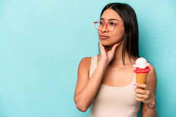 Young hispanic woman holding an ice cream isolated on blue background looking sideways with...