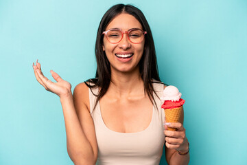Young hispanic woman holding an ice cream isolated on blue background receiving a pleasant...