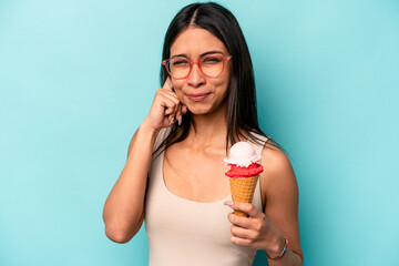 Young hispanic woman holding an ice cream isolated on blue background covering ears with hands.