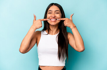 Young hispanic woman isolated on blue background smiles, pointing fingers at mouth.