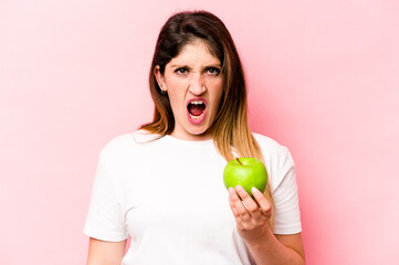 Young caucasian woman holding an apple isolated on pink background screaming very angry and aggressive.