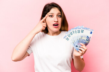 Young caucasian woman holding banknotes isolated on pink background showing a disappointment...