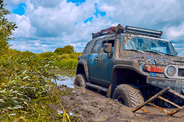 Obraz na płótnie Canvas Blue 4x4 car or 4WD car with wheels in mud on the off-road on a background of greenery and blue sky with clouds. An off-road car is stuck in a puddle of mud and is trying to leave using a ladder.