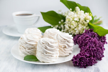Blurred image of marshmallows in a white plate, a cup of coffee and a bouquet of lilies of the valley and lilacs.