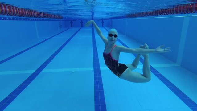 sportswoman of synchronized swimming in a tracksuit, in goggles for diving, in a cap for swimming is training in the pool. Gymnastic exercises and elements of somersaults in water. Wide angle, 4k