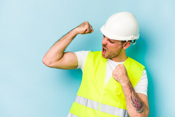 Young laborer caucasian man isolated on blue background raising fist after a victory, winner concept.