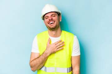 Young laborer caucasian man isolated on blue background laughs out loudly keeping hand on chest.