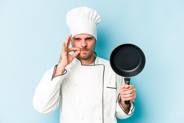 Young caucasian chef man holding flying pan isolated on blue background with fingers on lips keeping a secret.