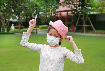 Asian little girl child wearing medical mask and straw hat with pointing one forefinger up due...