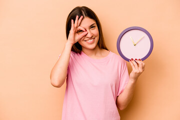 Young hispanic woman holding a clock isolated on beige background excited keeping ok gesture on eye.