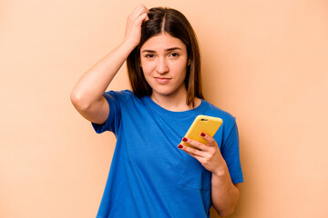 Young caucasian woman holding mobile phone isolated on beige background being shocked, she has...