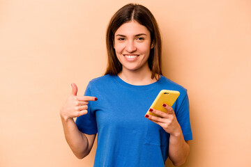 Young caucasian woman holding mobile phone isolated on beige background person pointing by hand to a shirt copy space, proud and confident