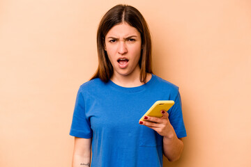 Young caucasian woman holding mobile phone isolated on beige background screaming very angry and...