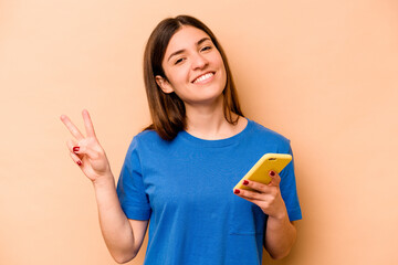 Young caucasian woman holding mobile phone isolated on beige background joyful and carefree showing a peace symbol with fingers.