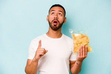 Young hispanic man holding a bag of chips isolated on blue background pointing upside with opened mouth.