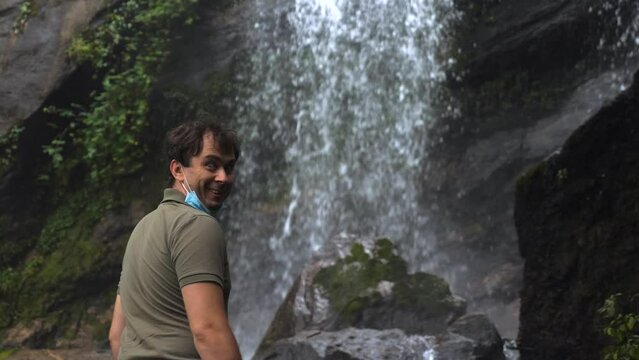 Happy funny travel man excited and enjoys a tropical waterfall. Smiling male tourist in face mask fooling around and fun on nature park