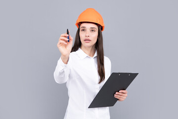 Serious woman in protective helmet with clipboard isolated on grey background. Young woman construction manager. Architect woman, female worker in hardhat helmet.
