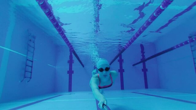 sportswoman swimmer in a tracksuit, in diving goggles, in a swimming cap is training in the pool. An athlete swims underwater with an action camera in her hand. Underwater shooting, wide angle, 4k